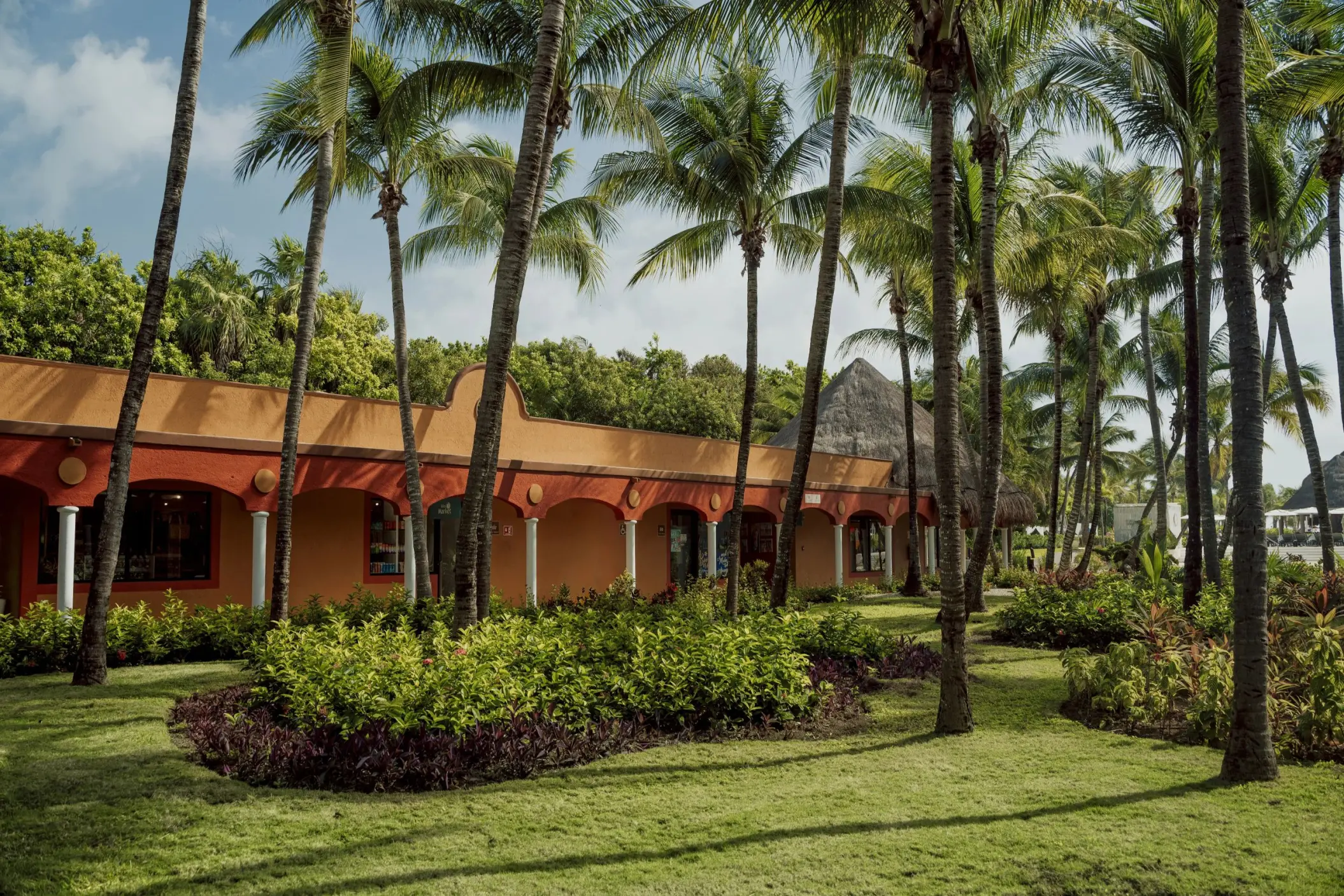 Lush green lawns and vibrant tropical plants surround a terracotta-colored resort building at Catalonia Playa Maroma, with tall palm trees.