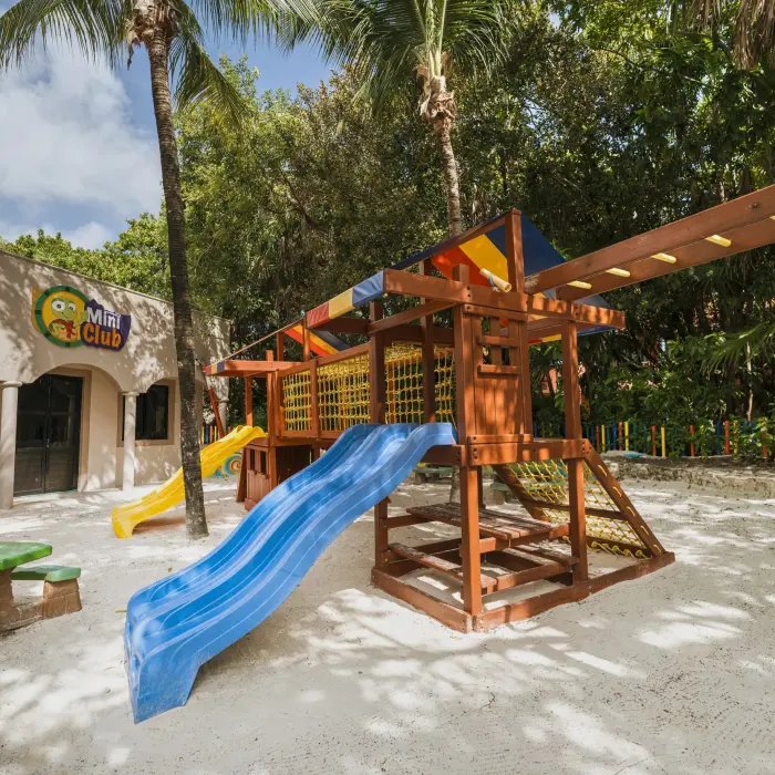A vibrant outdoor kids' play area at Catalonia Playa Maroma, featuring a wooden structure with slides, a sandpit, and a Mini Club building.