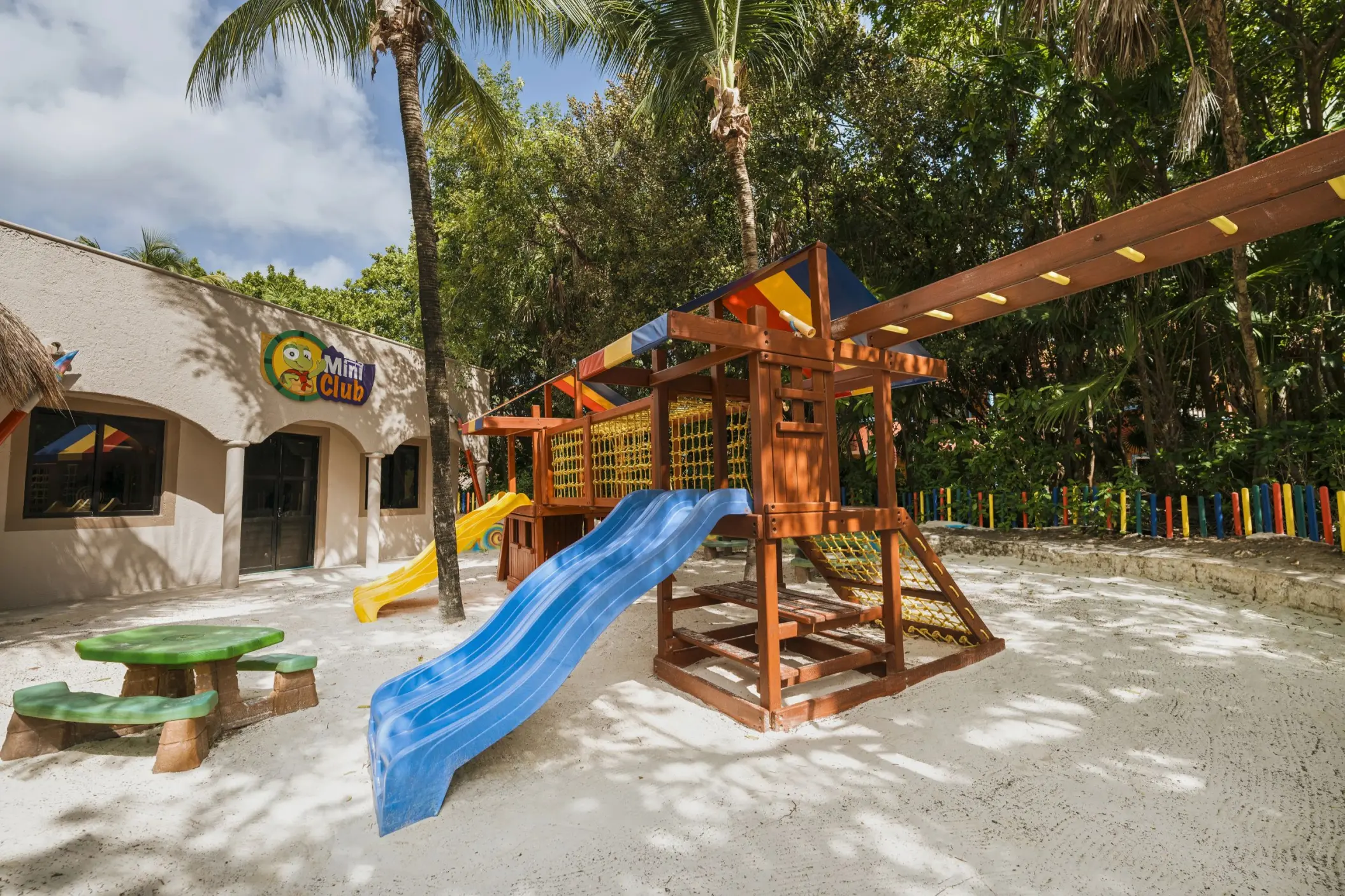 A vibrant outdoor kids' play area at Catalonia Playa Maroma, featuring a wooden structure with slides, a sandpit, and a Mini Club building.