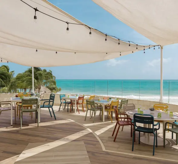 open-air ocean view restaurant at Catalonia Playa Maroma with a canvas shade, colorful tables and chairs, overlooking the turquoise sea.