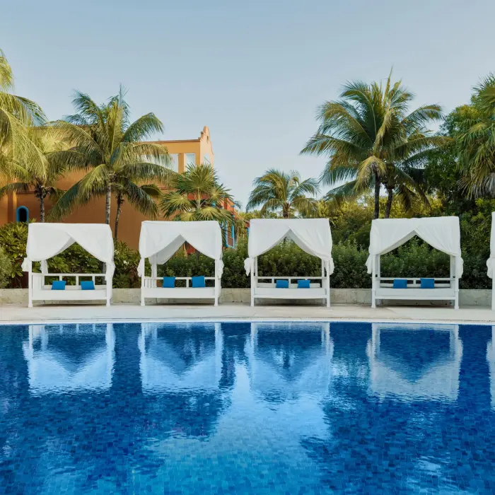 swimming pool at Catalonia Playa Maroma lined with elegant white Bali beds, framed by palm trees and resort buildings.