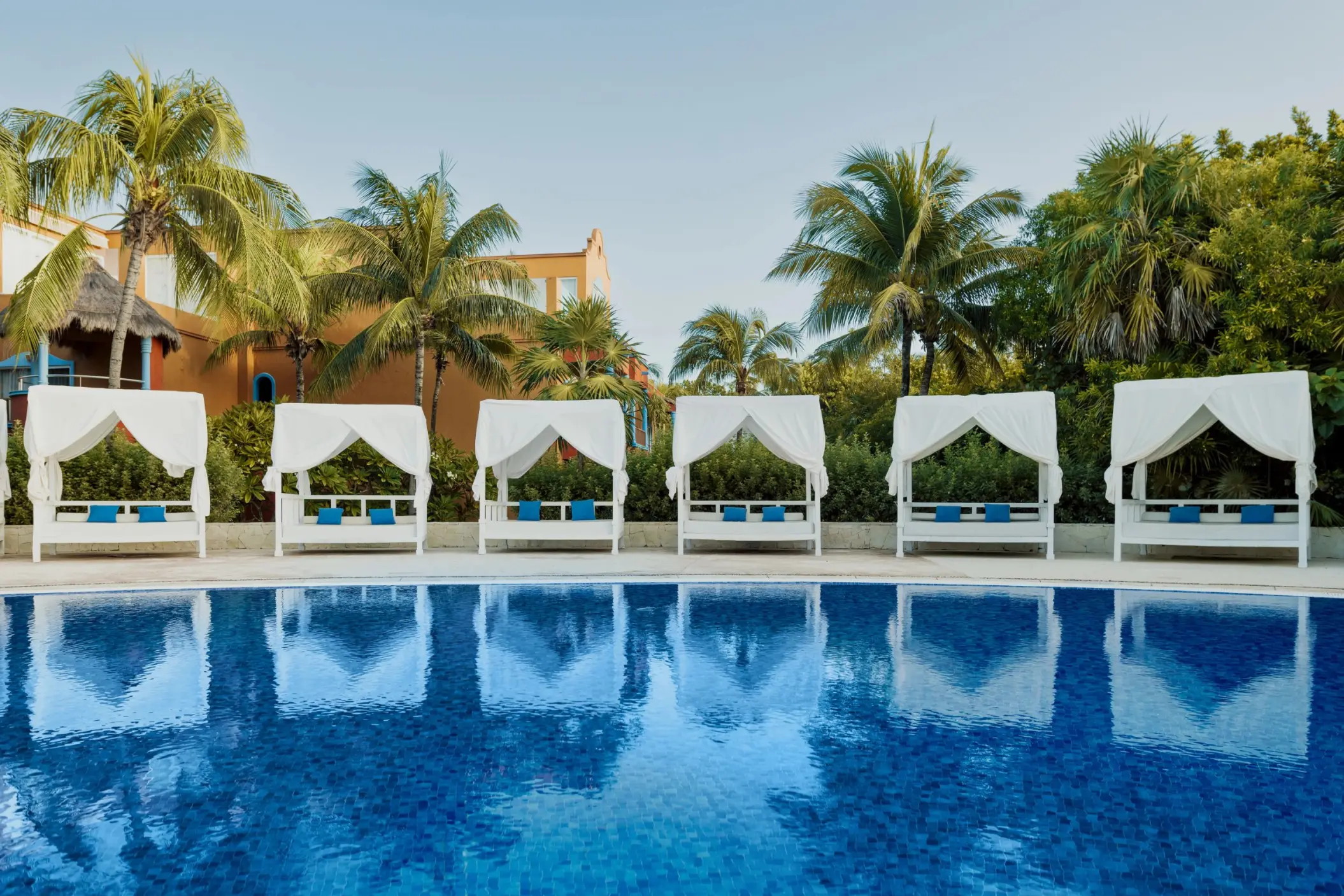 swimming pool at Catalonia Playa Maroma lined with elegant white Bali beds, framed by palm trees and resort buildings.