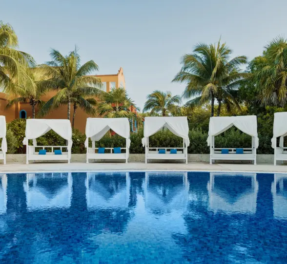 swimming pool at Catalonia Playa Maroma lined with elegant white Bali beds, framed by palm trees and resort buildings.