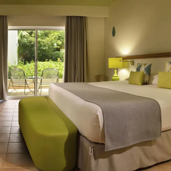 A cozy Privileged Superior suite at Catalonia Playa Maroma with a large bed, a green ottoman, and a sliding door leading to a balcony.