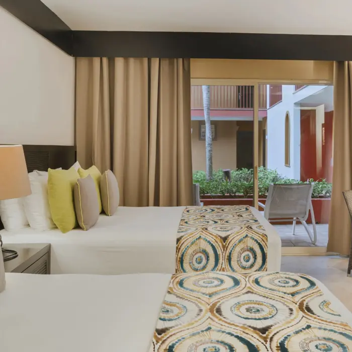 A comfortable resort room at Catalonia Playa Maroma with two beds, neutral tones, and a balcony view of the exterior.