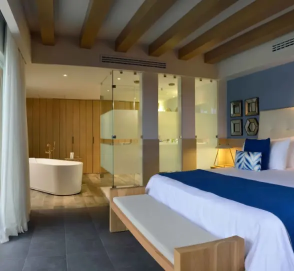 modern suite at Catalonia Playa Maroma with a large bed, blue accents, an open bathroom, and a balcony with a yellow chair.