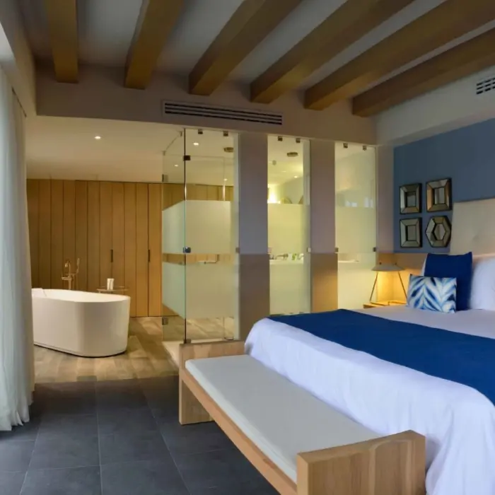 modern suite at Catalonia Playa Maroma with a large bed, blue accents, an open bathroom, and a balcony with a yellow chair.