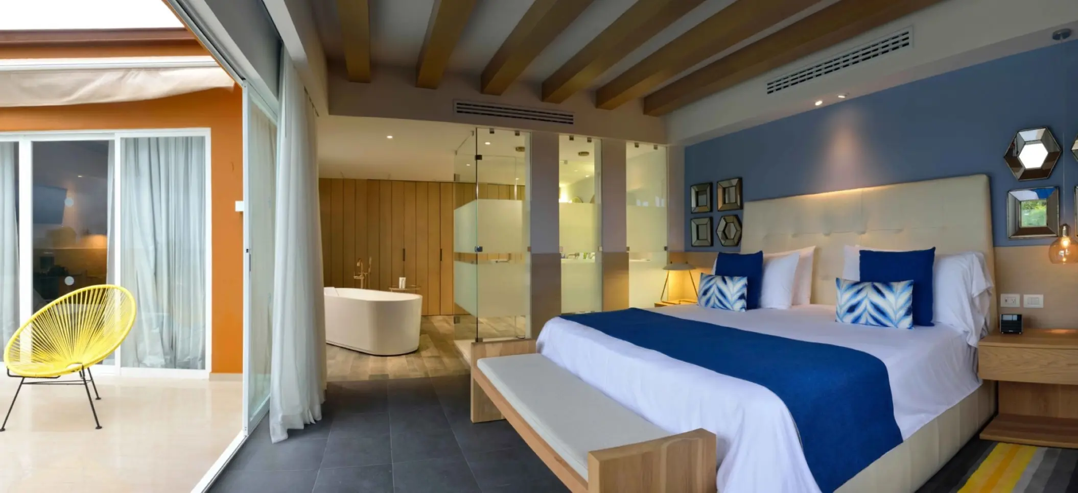 modern suite at Catalonia Playa Maroma with a large bed, blue accents, an open bathroom, and a balcony with a yellow chair.