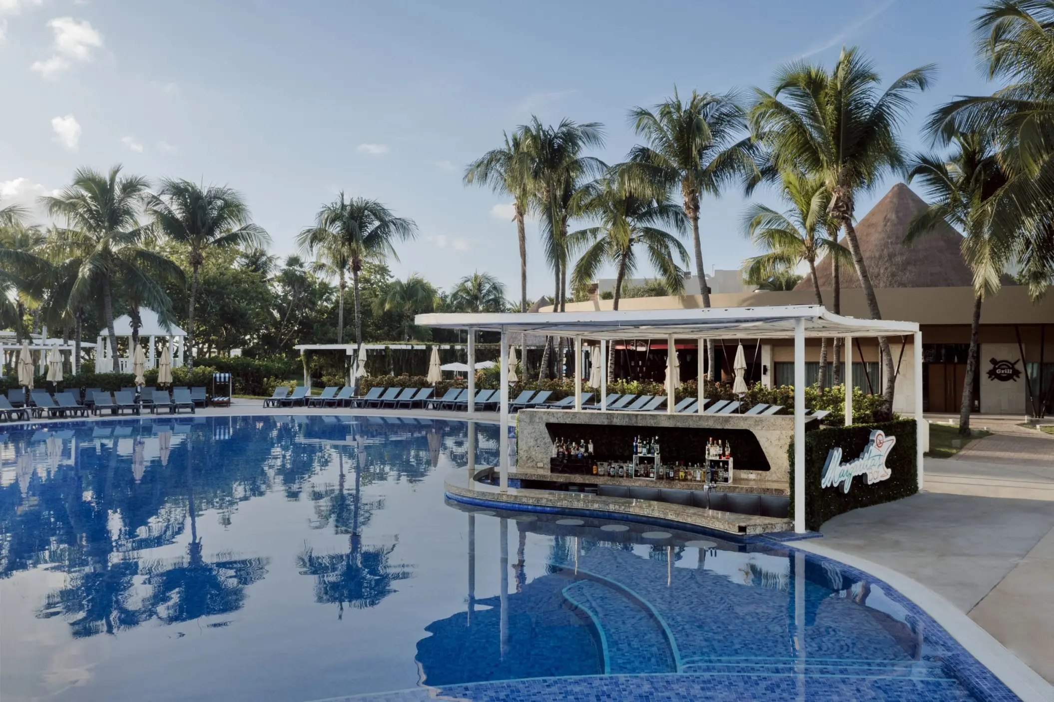 lively swim-up bar at Catalonia Playa Maroma, surrounded by a large pool, palm trees, and loungers, under a bright sky.