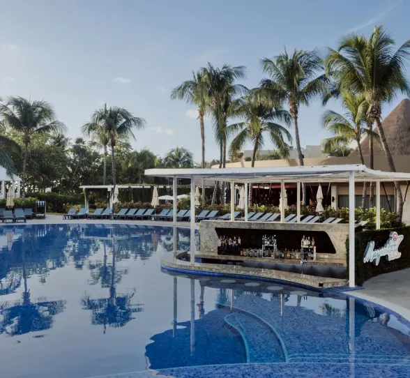lively swim-up bar at Catalonia Playa Maroma, surrounded by a large pool, palm trees, and loungers, under a bright sky.