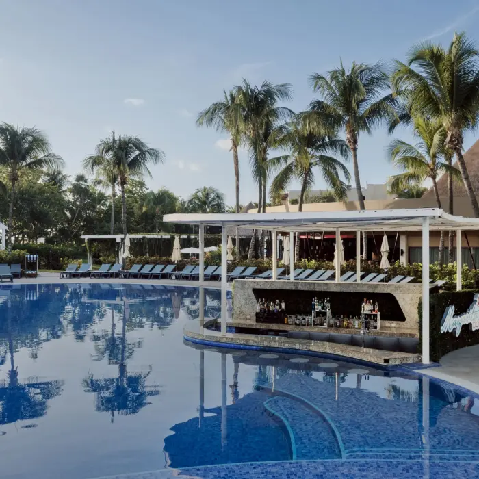 lively swim-up bar at Catalonia Playa Maroma, surrounded by a large pool, palm trees, and loungers, under a bright sky.