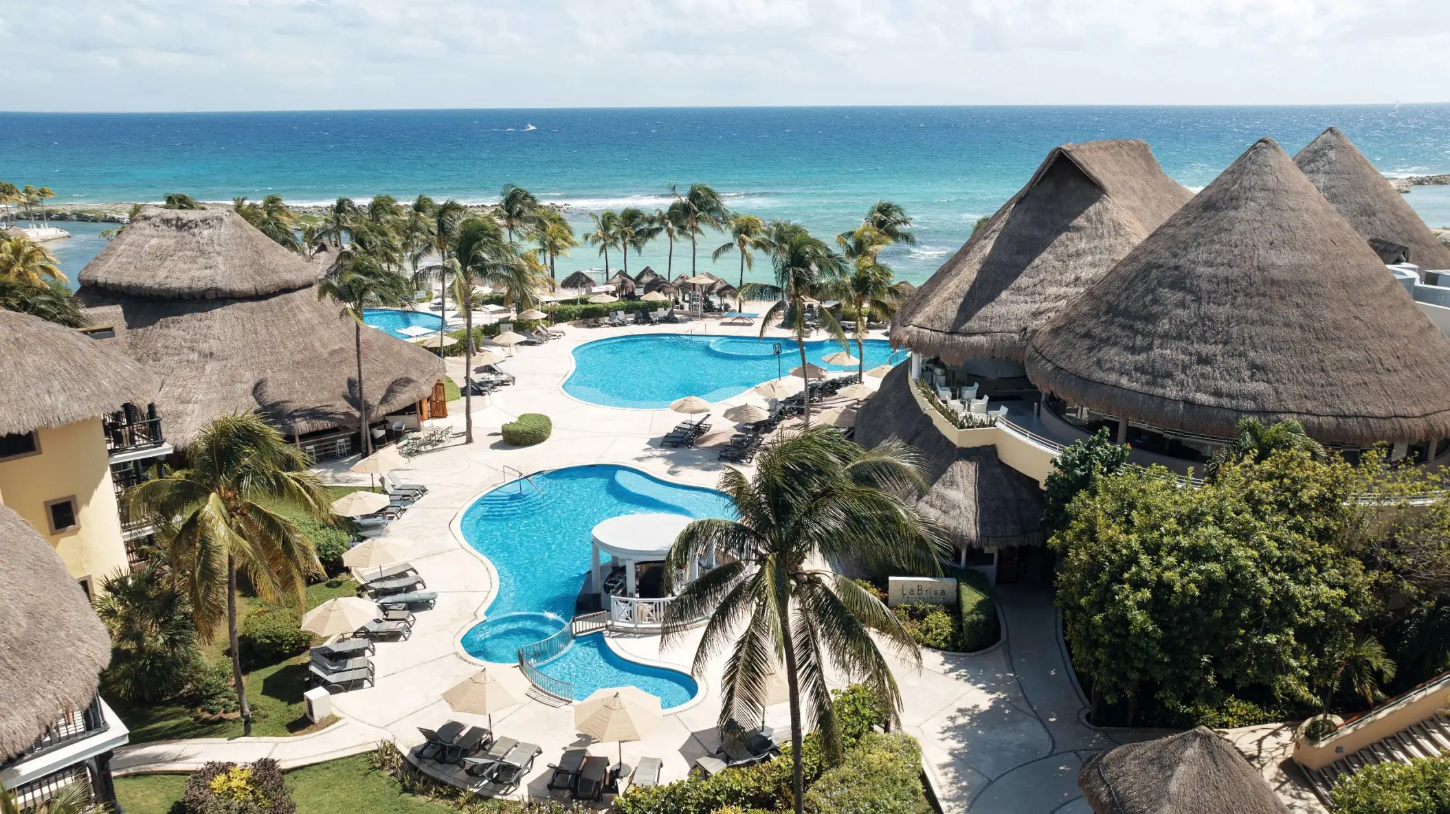 Aerial view of multiple turquoise pools and a thatched-roof swim-up bar, with the blue ocean in the background.
