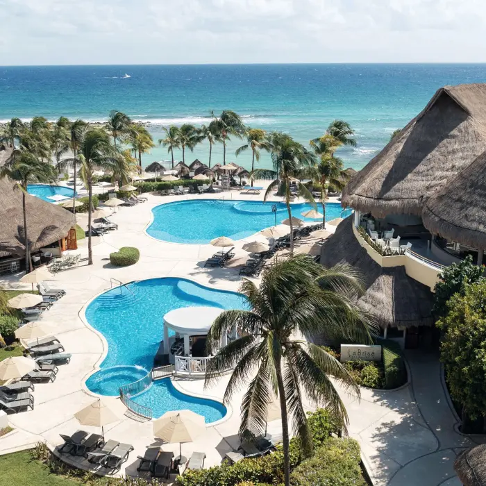 Aerial view of multiple turquoise pools and a thatched-roof swim-up bar, with the blue ocean in the background.