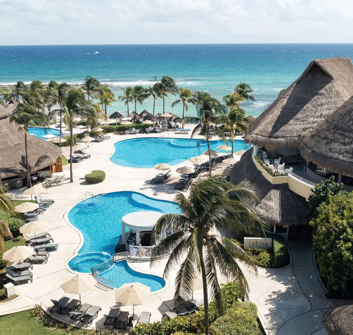 Aerial view of multiple turquoise pools and a thatched-roof swim-up bar, with the blue ocean in the background.