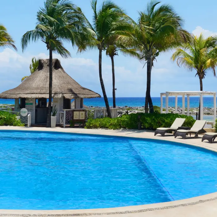 A bright blue swimming pool surrounded by palm trees, lounge chairs, and a small hut, with the ocean in the background.