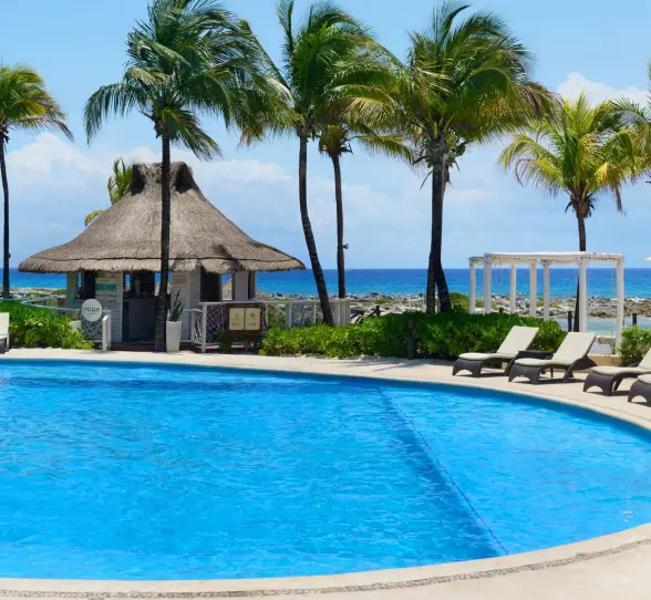 A bright blue swimming pool surrounded by palm trees, lounge chairs, and a small hut, with the ocean in the background.