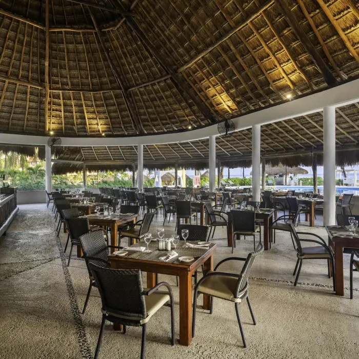 A spacious open-air restaurant with a thatched roof, set up with multiple tables and chairs near the pool.