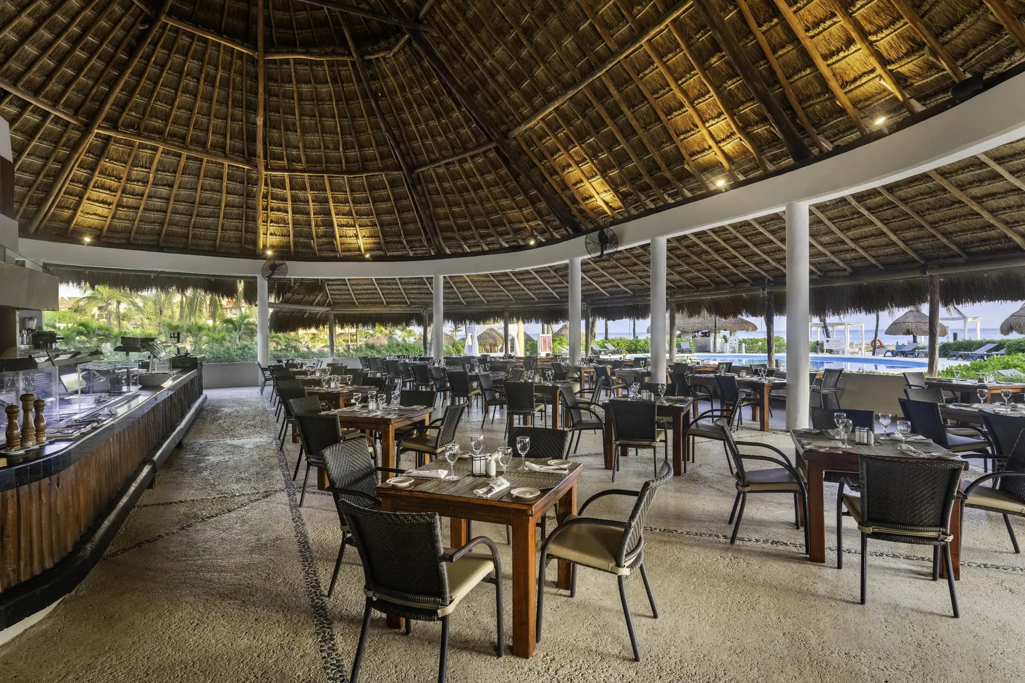 A spacious open-air restaurant with a thatched roof, set up with multiple tables and chairs near the pool.