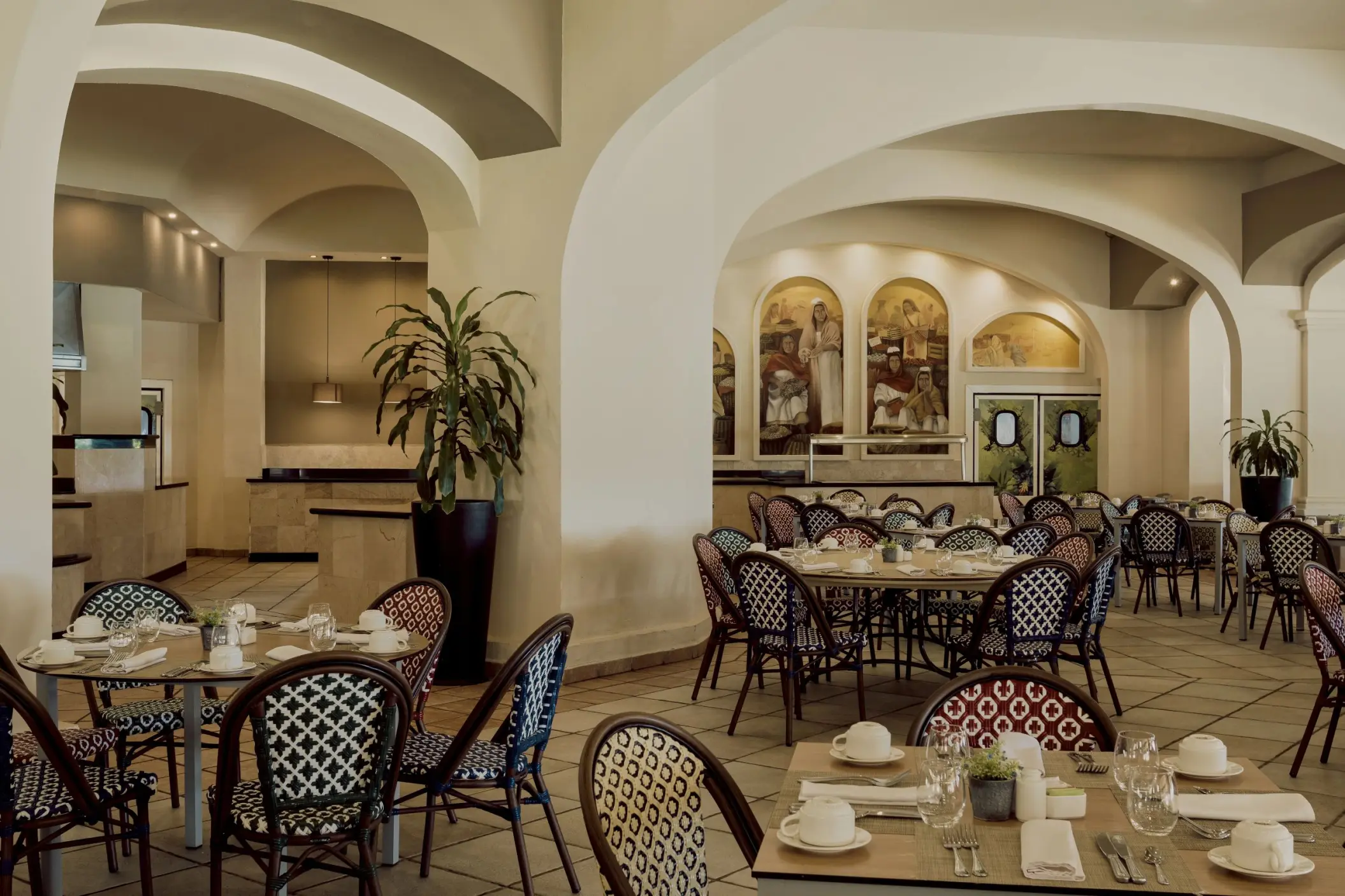 An elegant restaurant with arched doorways, round tables with decorative chairs, and murals on the walls.