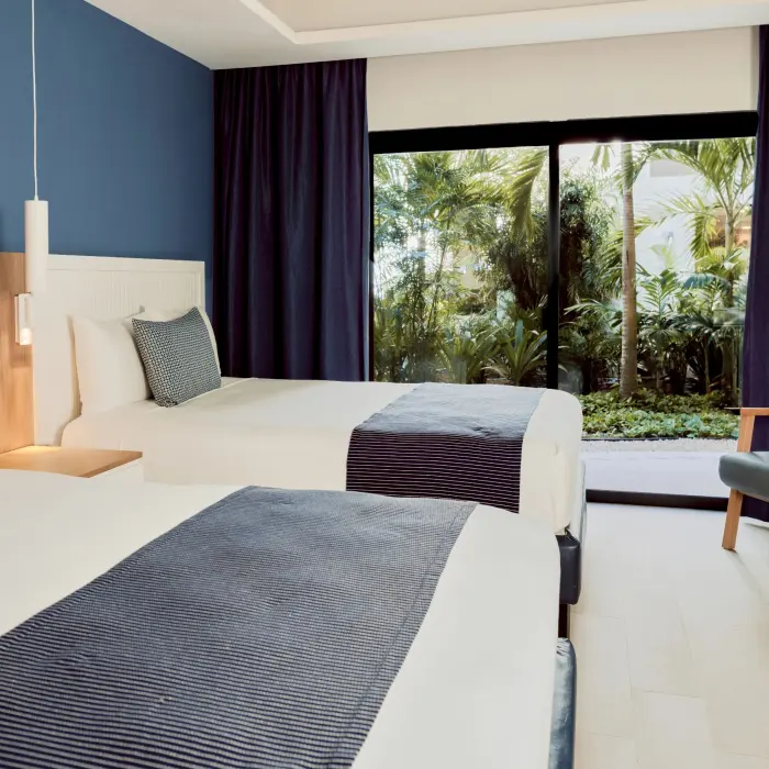 A hotel room featuring two beds with blue accents, a blue accent wall, and a view of lush greenery outside.