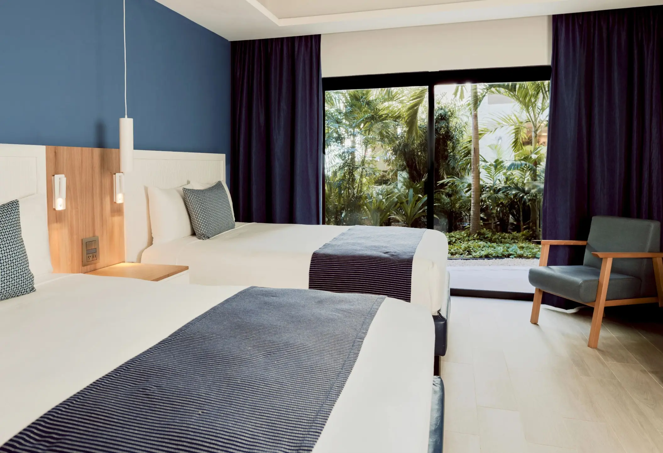 A hotel room featuring two beds with blue accents, a blue accent wall, and a view of lush greenery outside.