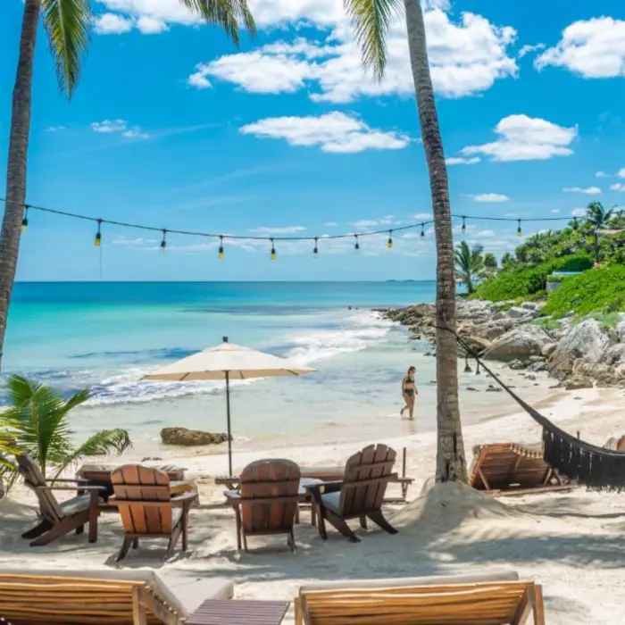 A picturesque white sand beach with turquoise water, palm trees, hammocks, and string lights.