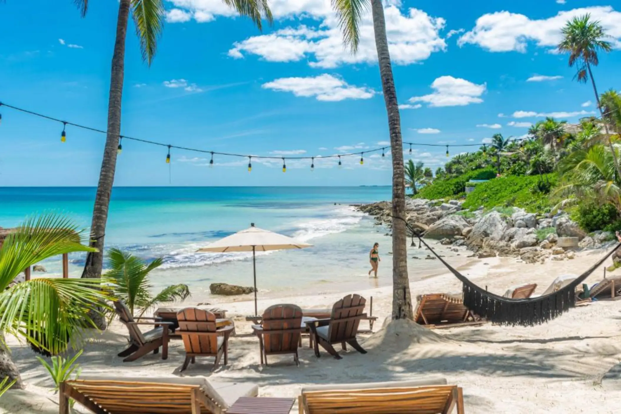 A picturesque white sand beach with turquoise water, palm trees, hammocks, and string lights.