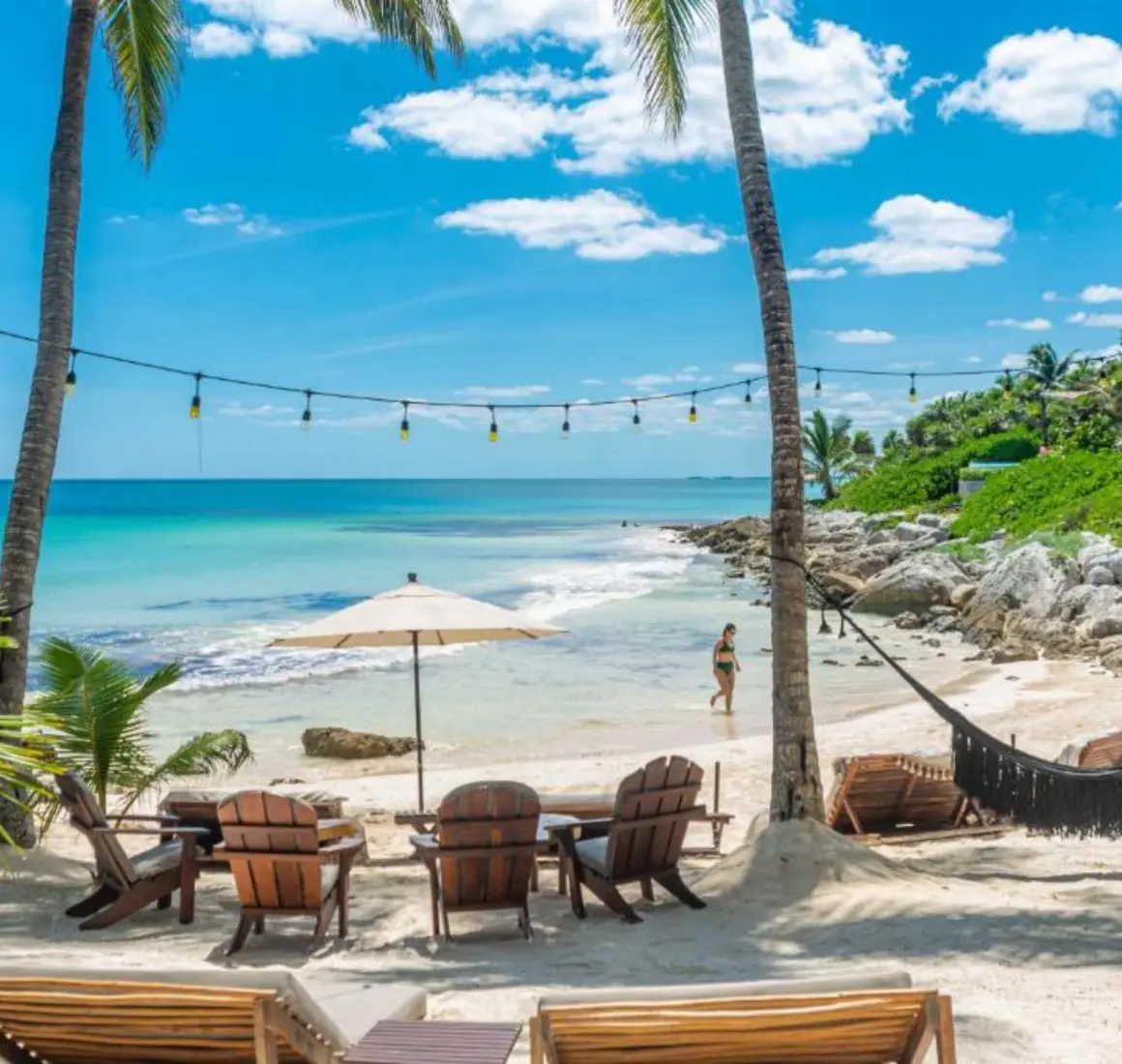A picturesque white sand beach with turquoise water, palm trees, hammocks, and string lights.