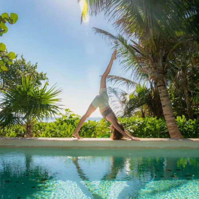 A person doing yoga by a serene pool, surrounded by vibrant green tropical foliage and ocean in distance.