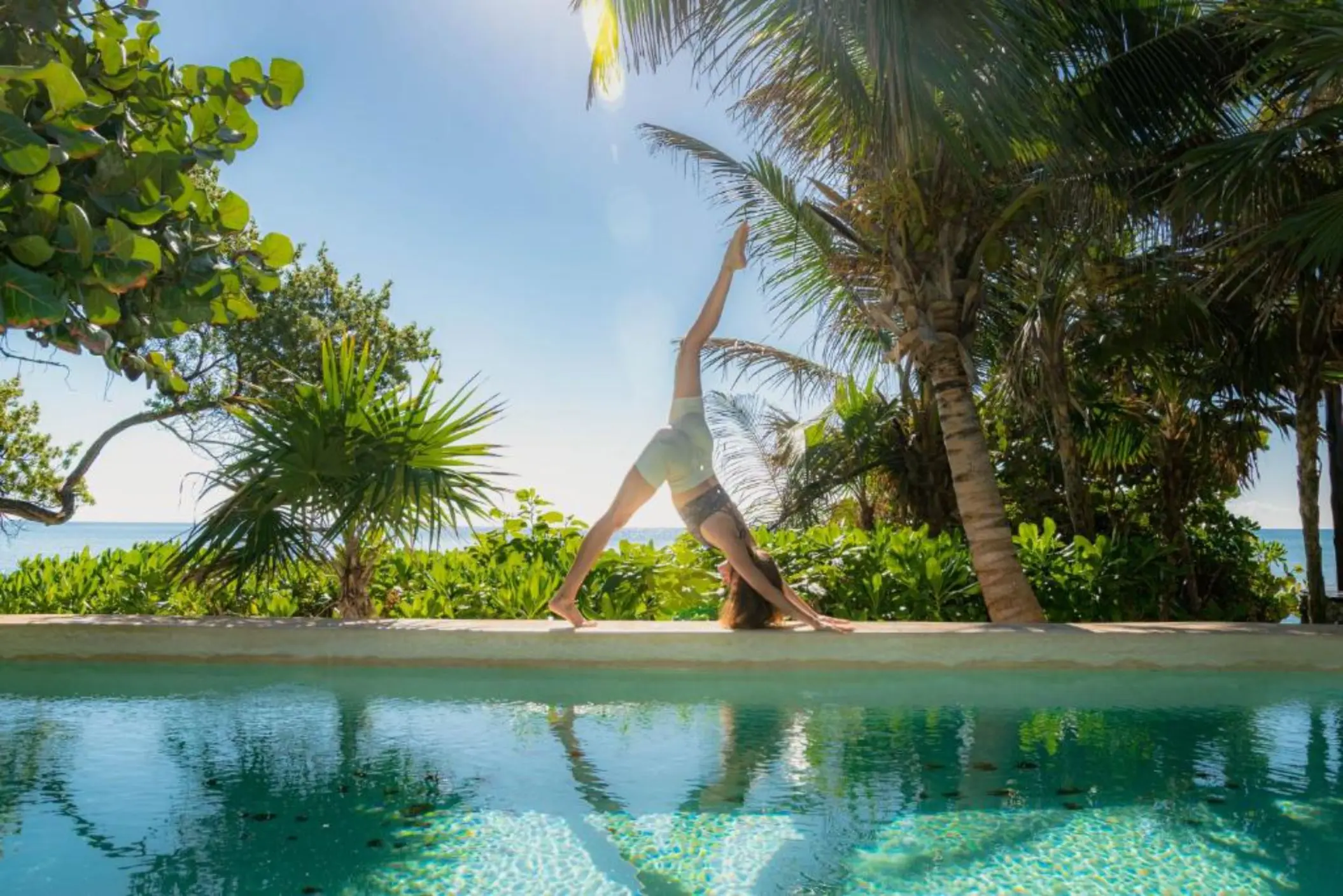 A person doing yoga by a serene pool, surrounded by vibrant green tropical foliage and ocean in distance.