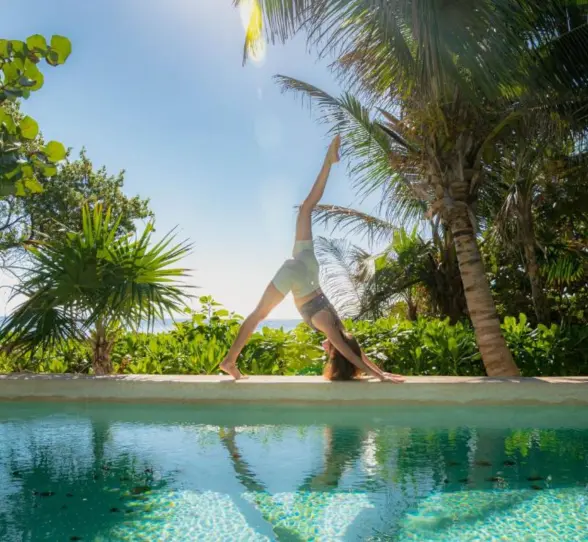 A person doing yoga by a serene pool, surrounded by vibrant green tropical foliage and ocean in distance.