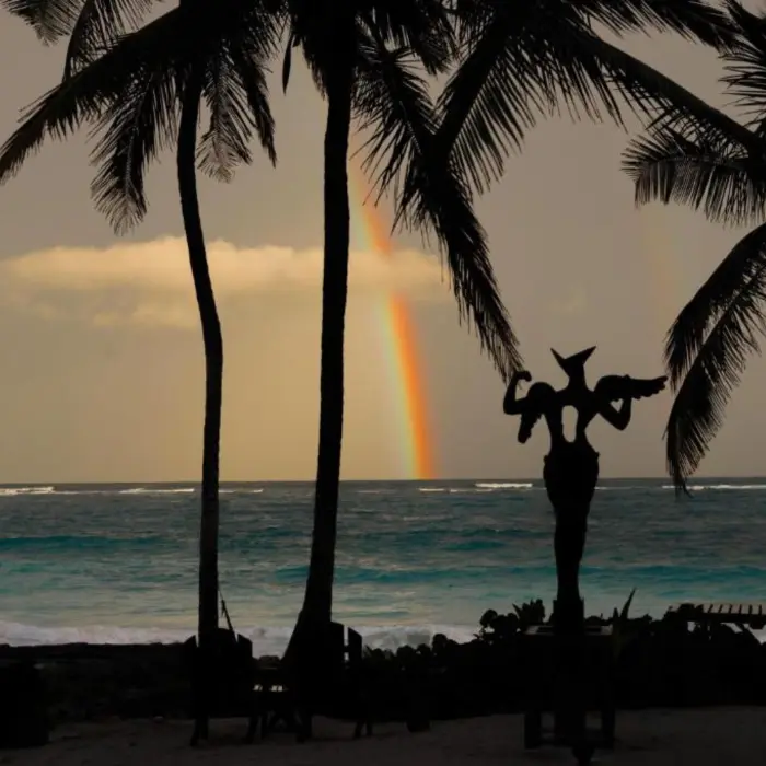 A rainbow arches over the ocean, with an artistic sculpture silhouetted against the sky.