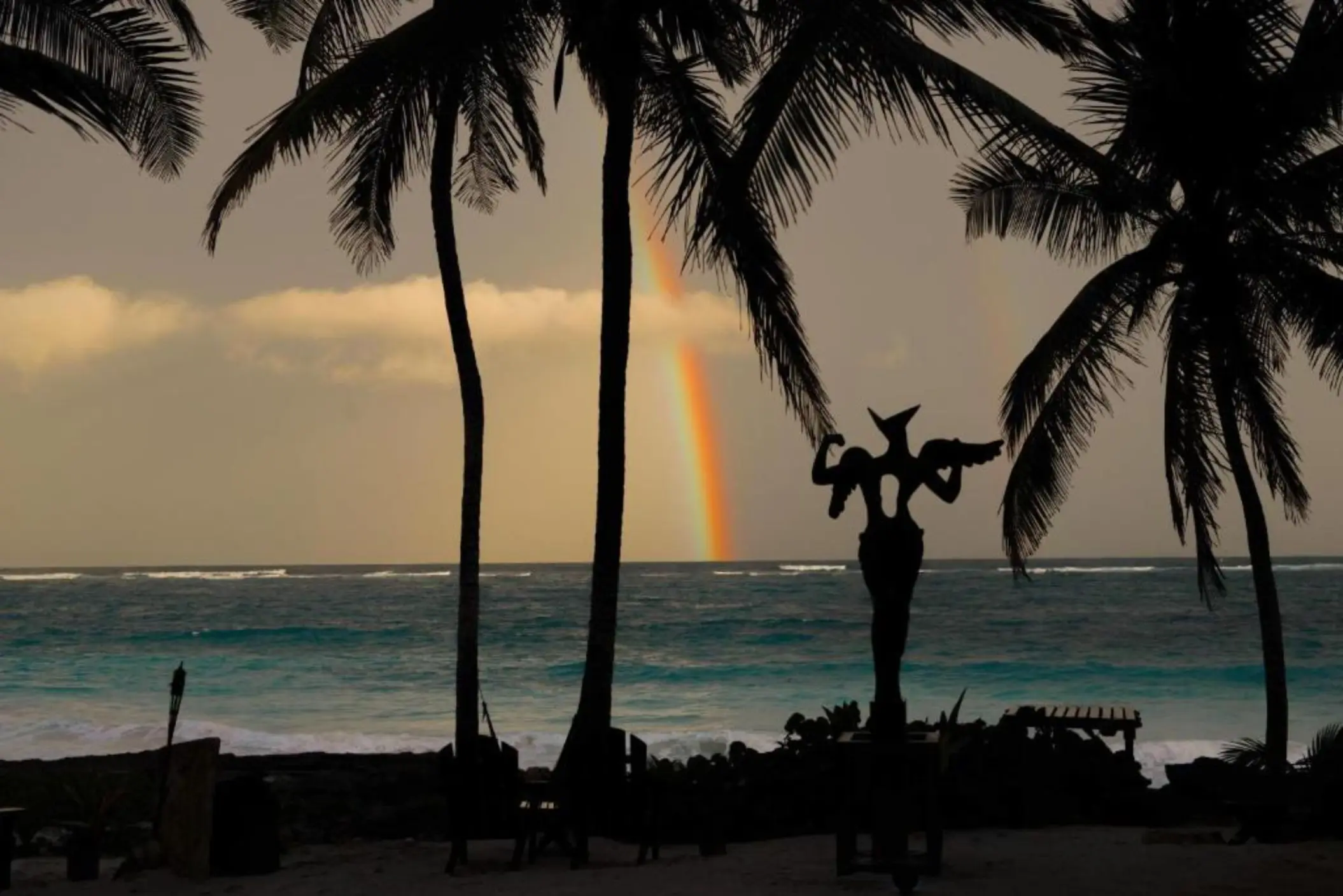 A rainbow arches over the ocean, with an artistic sculpture silhouetted against the sky.