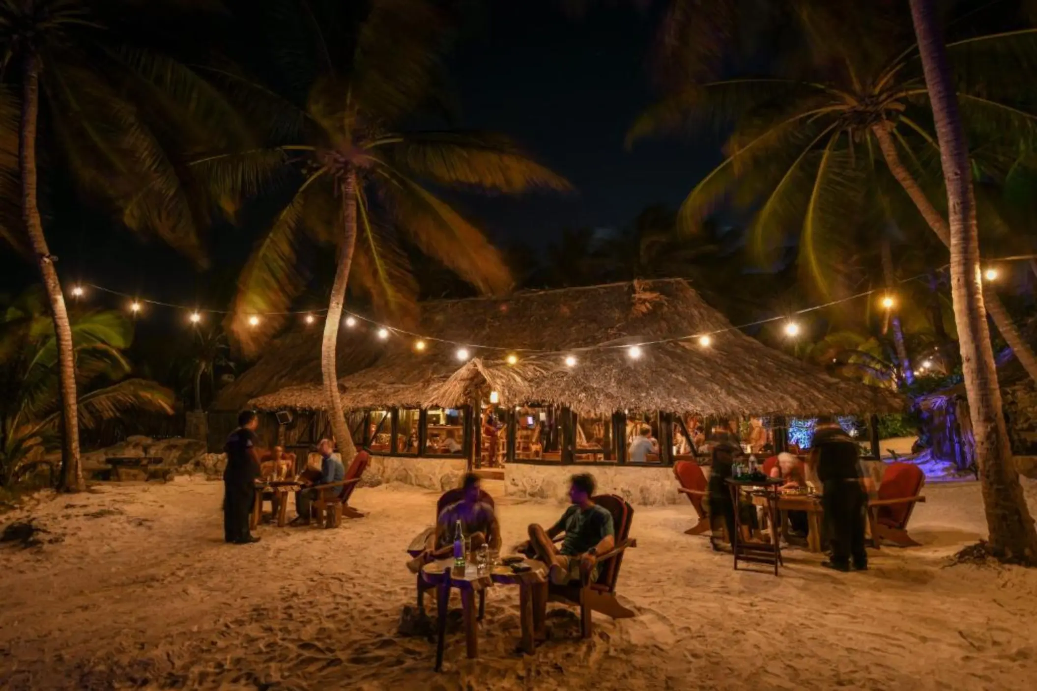 A charming open-air restaurant at night, illuminated by string lights, with guests dining on the sand.