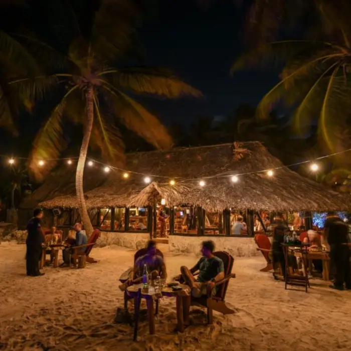 A charming open-air restaurant at night, illuminated by string lights, with guests dining on the sand.