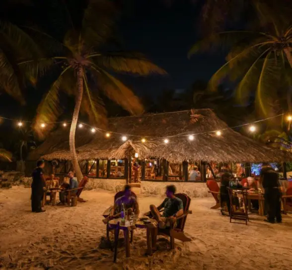A charming open-air restaurant at night, illuminated by string lights, with guests dining on the sand.