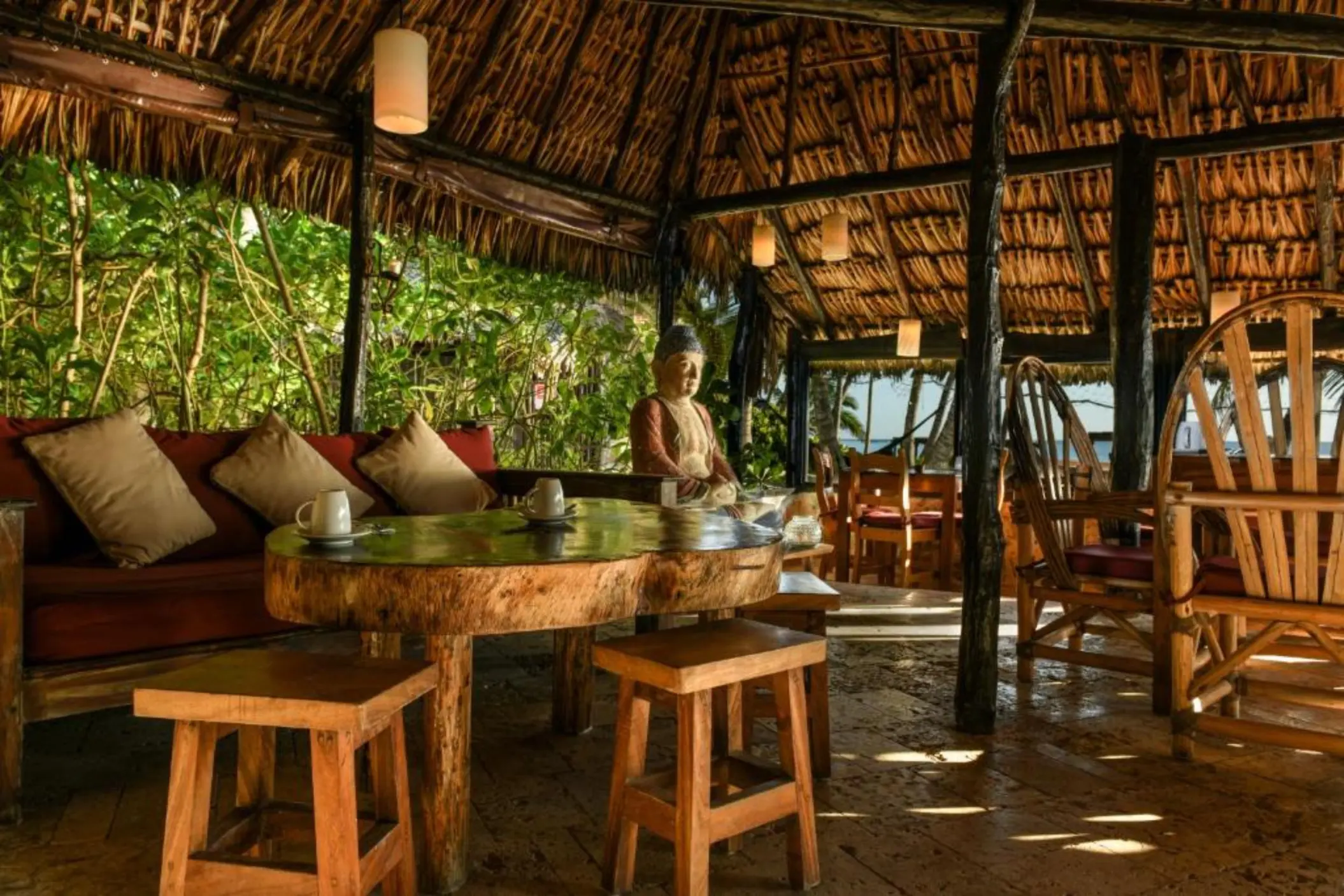 The rustic interior of a restaurant with a thatched roof, wooden furniture, and a Buddha statue.