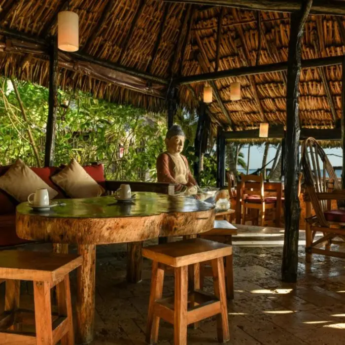 The rustic interior of a restaurant with a thatched roof, wooden furniture, and a Buddha statue.