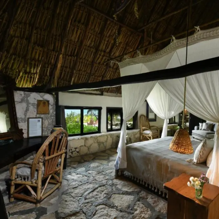 A rustic-chic suite with a canopy king bed, stone floor, and ocean view through large windows.