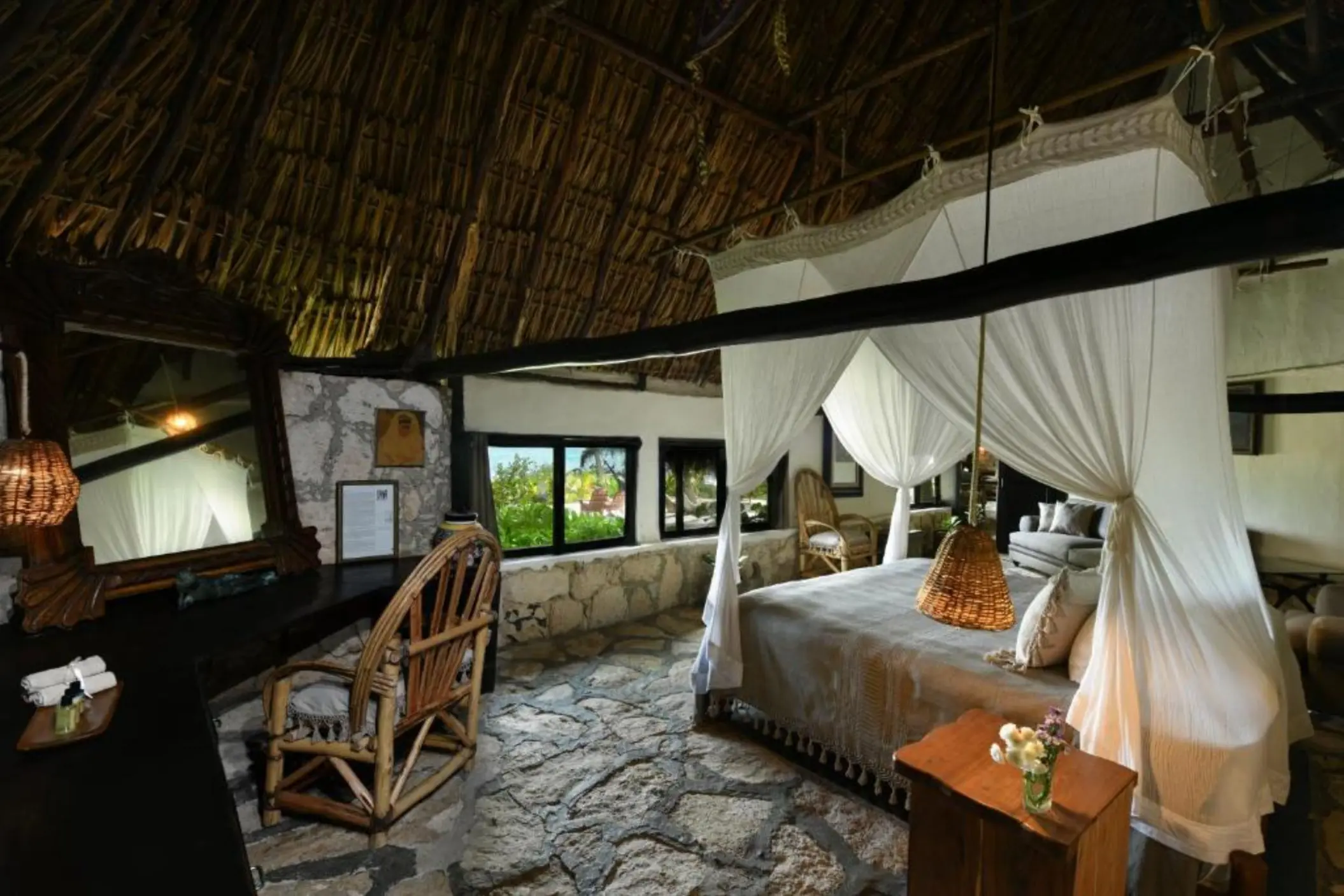 A rustic-chic suite with a canopy king bed, stone floor, and ocean view through large windows.