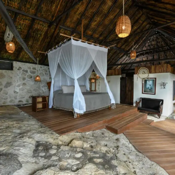 The interior of a unique suite with a canopy king bed, thatched roof, and natural stone and wood elements.