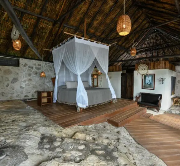 The interior of a unique suite with a canopy king bed, thatched roof, and natural stone and wood elements.