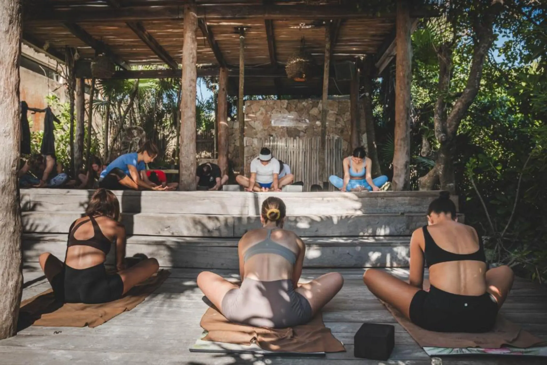 A group of people practicing yoga outdoors under a rustic, open-air pavilion surrounded by trees.
