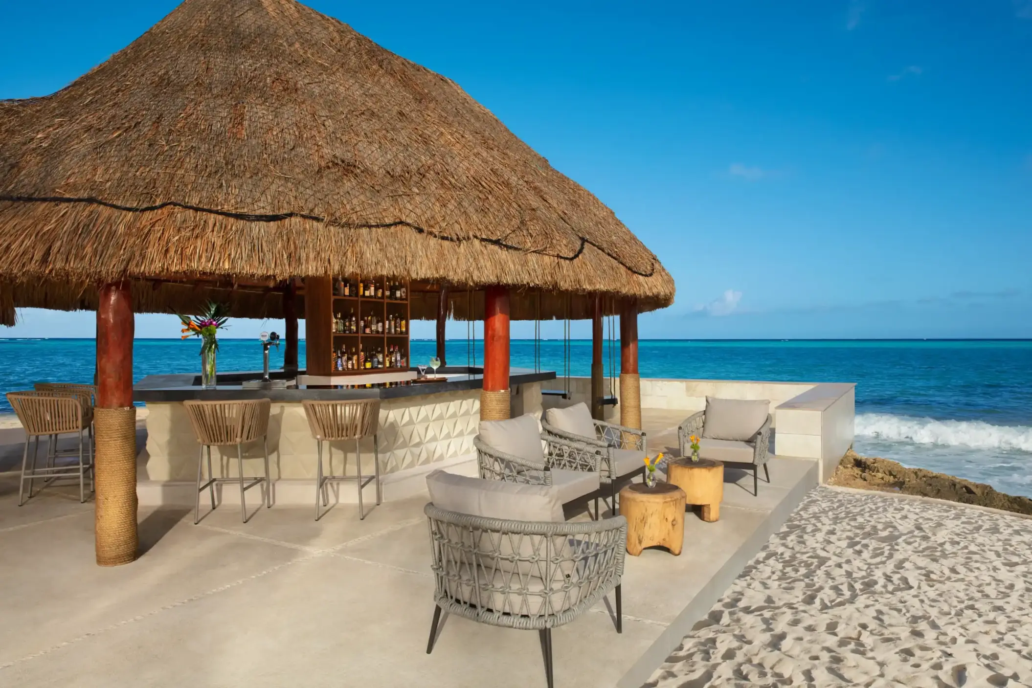 Oceanfront beach bar with lounge chairs and umbrellas on white sand, next to a palapa-style structure by the sea.