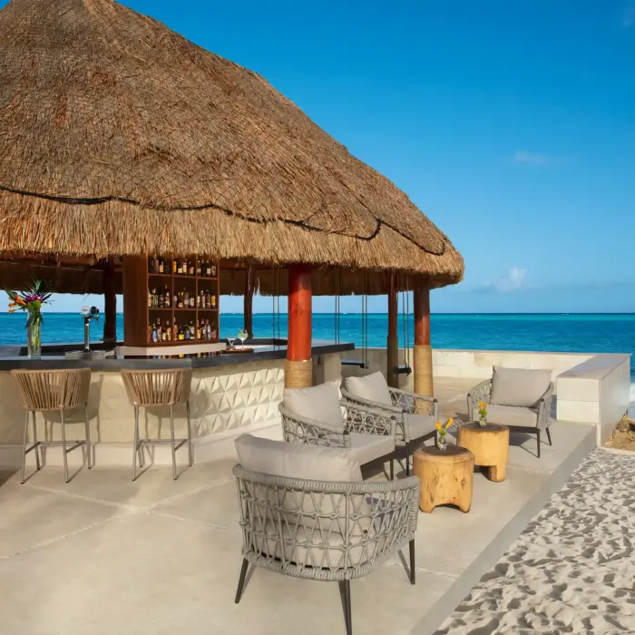 Oceanfront beach bar with lounge chairs and umbrellas on white sand, next to a palapa-style structure by the sea.
