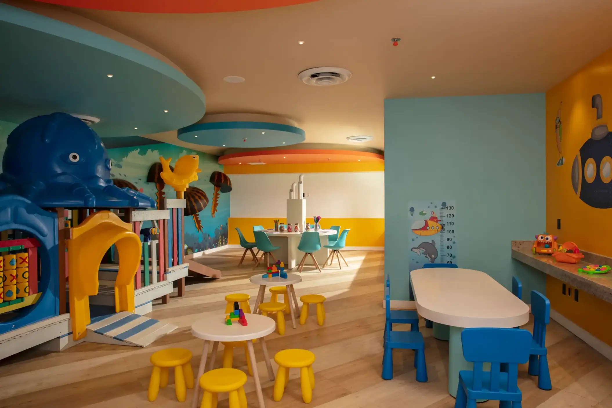 Bright indoor kids' play area with colorful tables, ocean-themed walls, and a soft structure with sea animal elements.