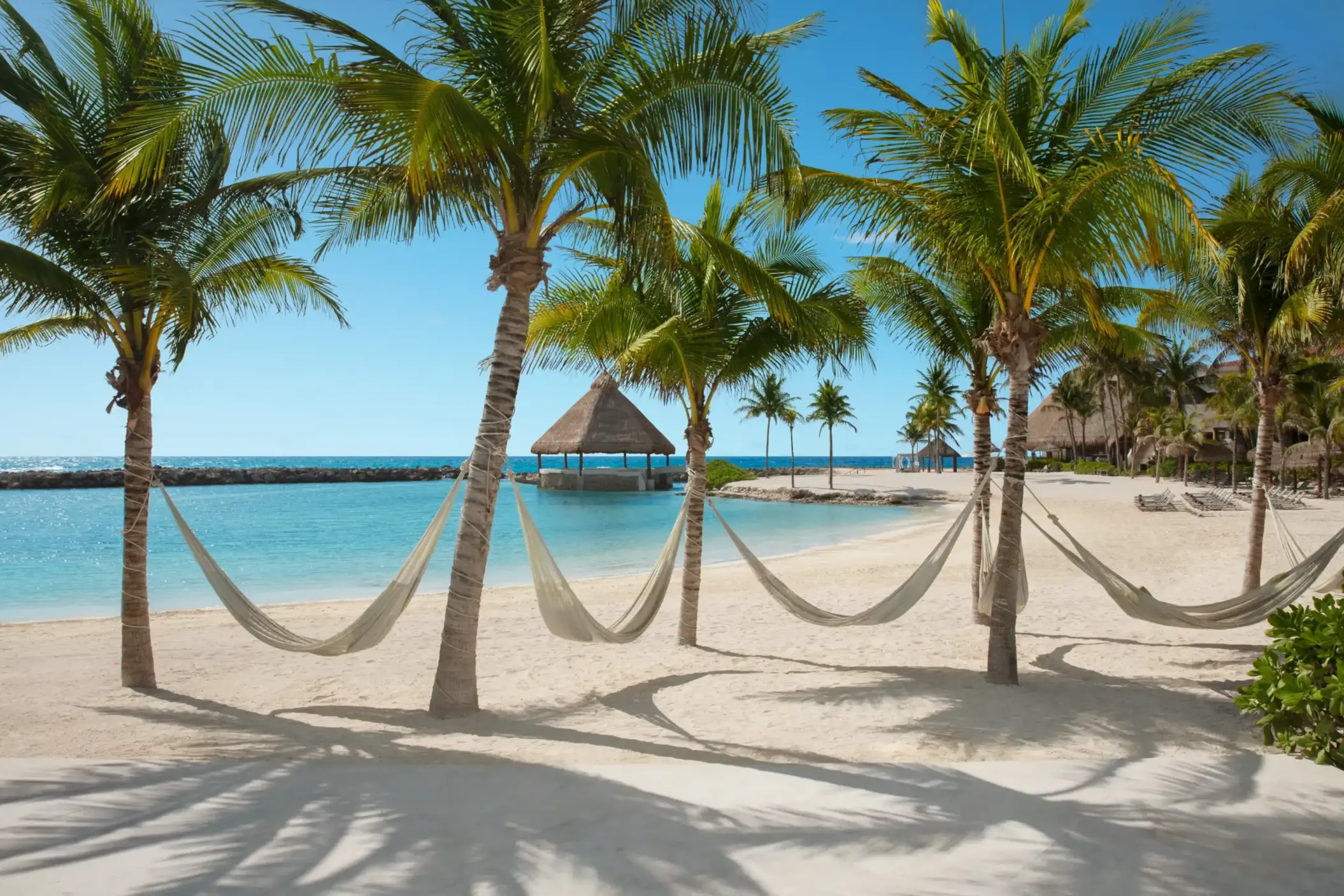 Tropical beach with hammocks strung between palm trees, thatched pier over turquoise water, white sand resort.
