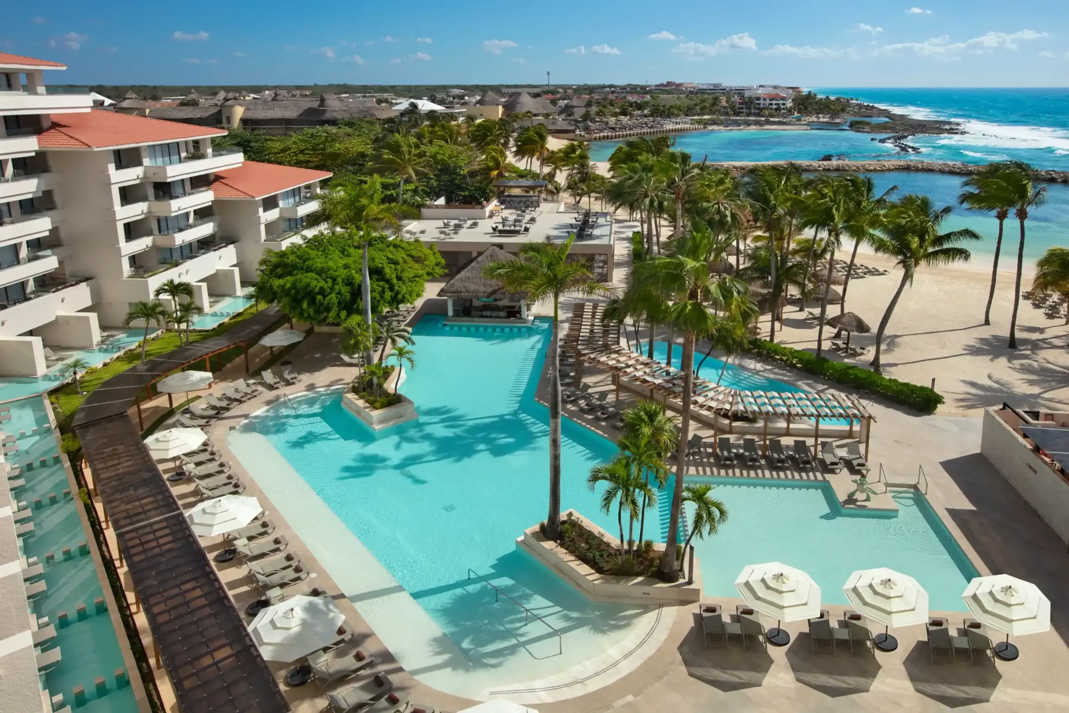 Expansive resort pool with sun loungers, palm trees, shaded walkways, and beach access overlooking crystal-clear waters.
