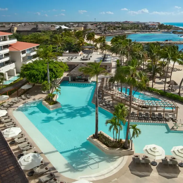 Expansive resort pool with sun loungers, palm trees, shaded walkways, and beach access overlooking crystal-clear waters.