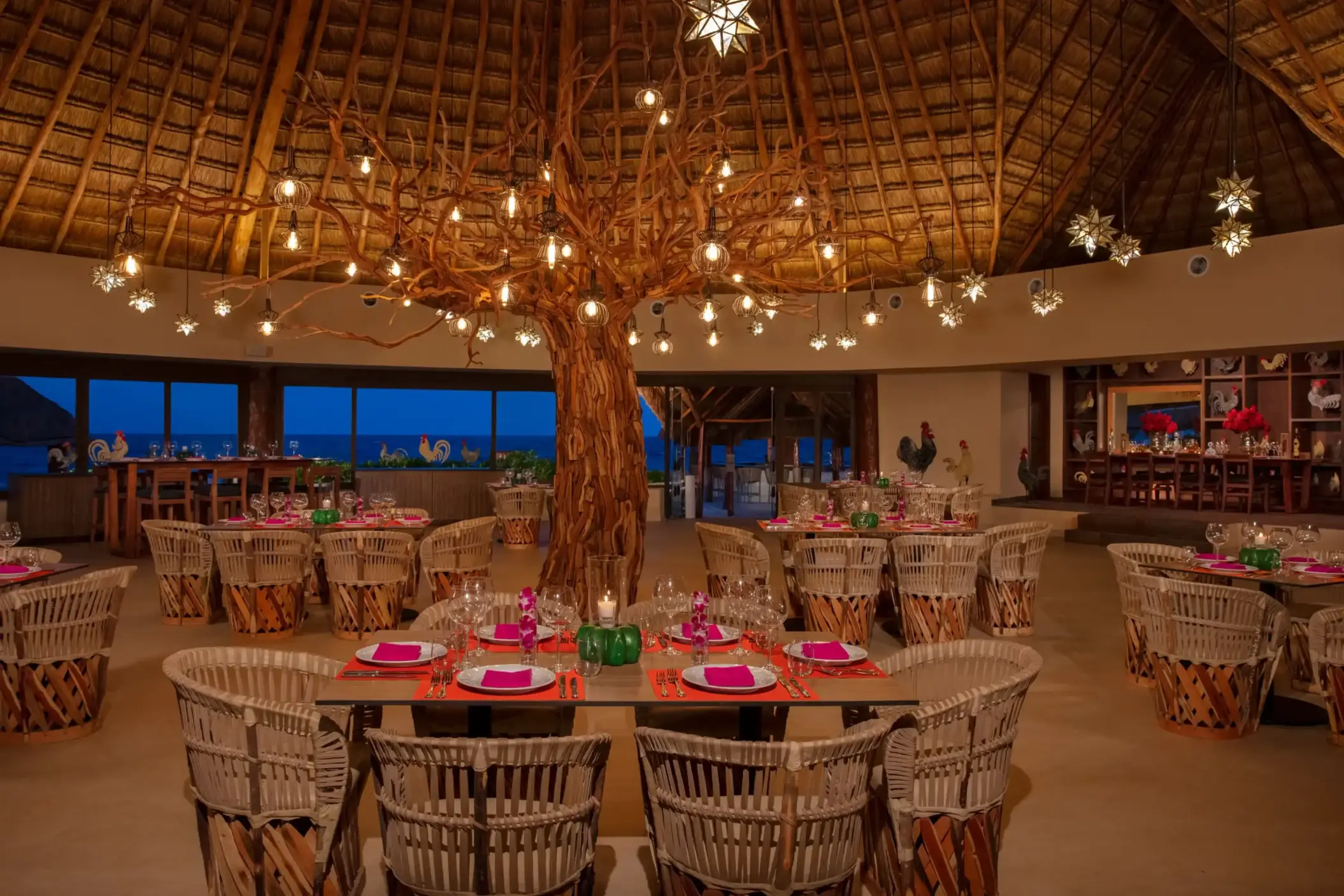 Rustic-style restaurant with a large tree centerpiece, woven chairs, and ambient lighting under a palapa roof.